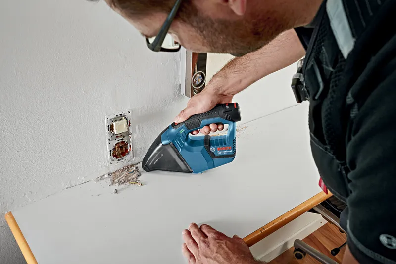 GAS 12 V-LI A person wearing safety equipment cleans debris near an electrical outlet with a cordless vacuum.
