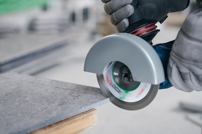 A person wearing safety equipment cuts a tile with an angle grinder.