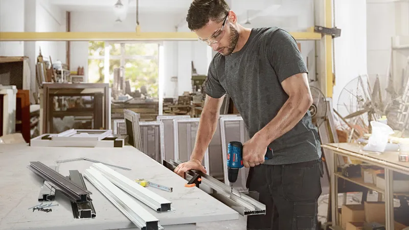 A person wearing safety equipment drills into a metal profile at a workbench.