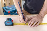 Person marking a measurement on wood with a pencil using a tape measure.