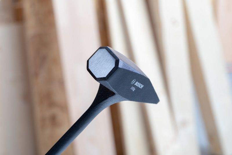 Black sledgehammer head with a flat striking face, shown against a blurred wood background.