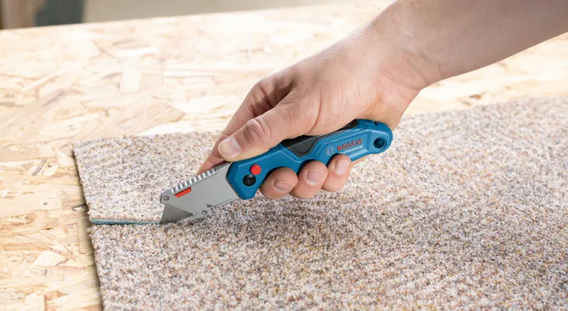 A person uses a folding knife to cut through a piece of carpet.