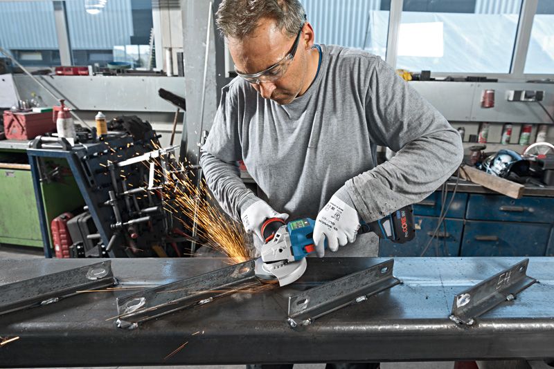 A person wearing safety equipment uses an angle grinder to cut a metal beam.
