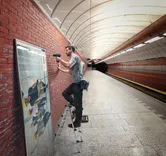A person using a cordless drill to mount a frame on a brick wall in a subway station.