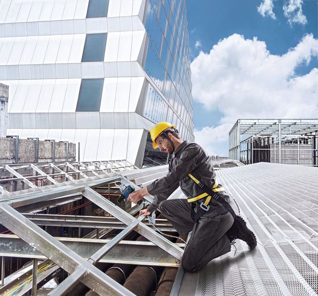 A person wearing safety equipment drills into a metal frame on a rooftop construction site.