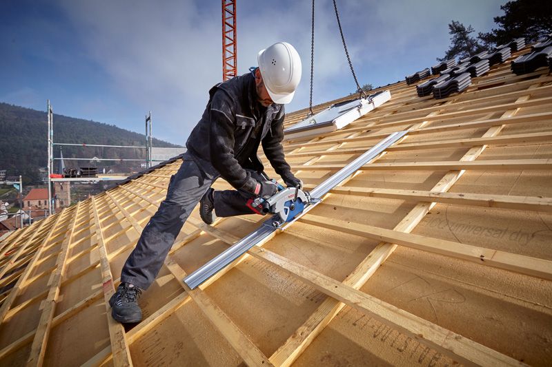 A person wearing safety equipment cuts wood on a sloped roof using a track saw.