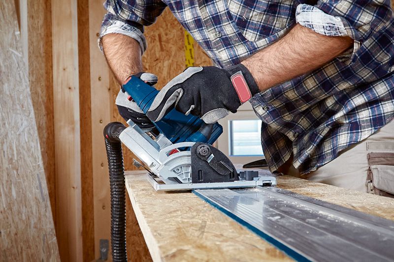 A person wearing safety equipment uses a circular saw to cut wood along a guide rail.