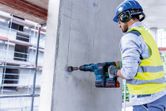 Person wearing safety equipment drills into a concrete wall with a cordless rotary hammer.
