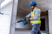 A person wearing safety equipment uses a cordless rotary hammer to drill into a concrete wall.