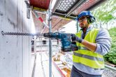 A person wearing safety equipment drills into concrete using a cordless rotary hammer.