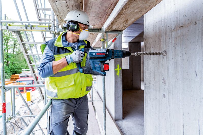 A person wearing safety equipment drills into concrete with a cordless rotary hammer.