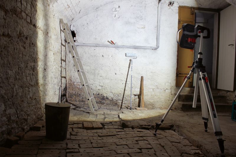 Cordless jobsite light on a tripod illuminates a basement with a ladder and painting tools.