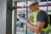 A person wearing safety equipment fastens a window frame with a cordless drill.