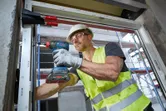 A person wearing safety equipment drives a screw into a window frame with a cordless drill.