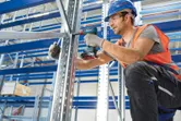 A person wearing safety equipment fastens a metal frame using a cordless power drill.