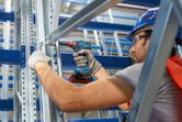 A person wearing safety equipment uses a cordless drill on a metal frame.