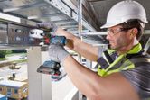A person wearing safety equipment uses a cordless drill on a steel beam.
