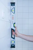 Person holding a 60cm optical level upright against a tiled wall.
