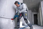A person wearing safety equipment drills into a concrete wall in a construction area.