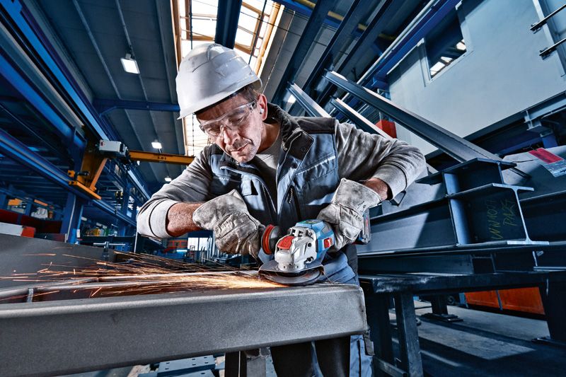 A person wearing safety equipment grinds metal with a cordless angle grinder, producing sparks.