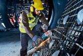A person wearing safety equipment uses a cordless angle grinder to cut metal rebar.