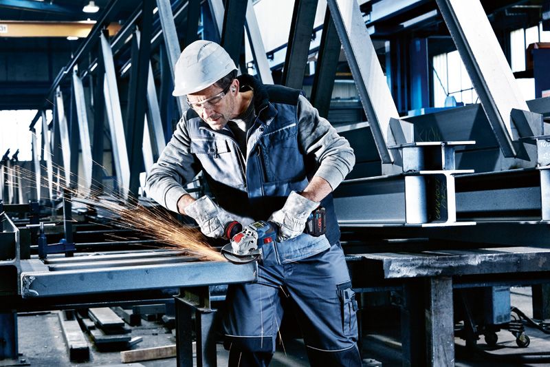 A person wearing safety equipment grinds metal beams with a cordless angle grinder.