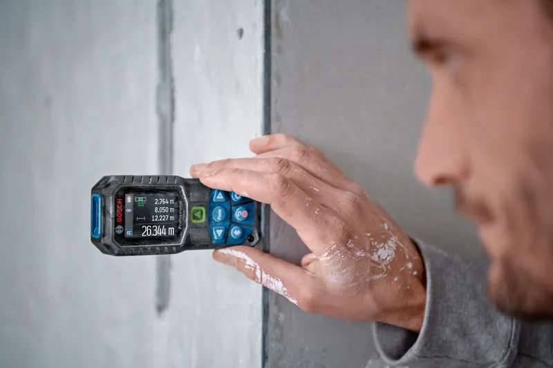 GLM 50-27 CG Person measures a wall with a laser measure, paint visible on their hand.