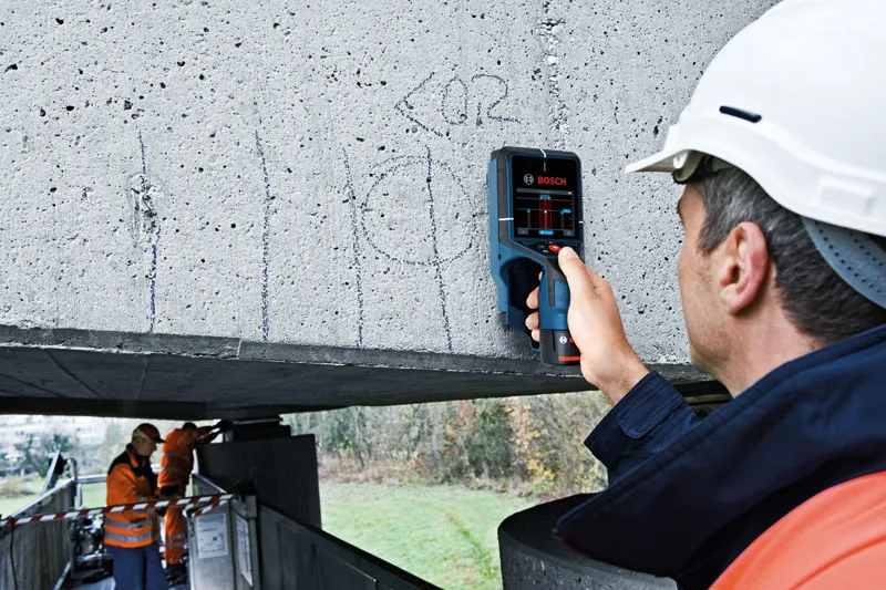 A person wearing safety equipment scans a concrete beam with an electronic detector.