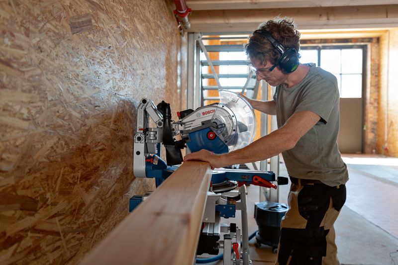 GCM 18V-305 GDC A person wearing safety equipment cuts a wooden beam using a cordless mitre saw.