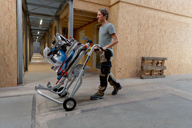 A person wearing safety equipment moves a cordless mitre saw on a wheeled stand indoors.