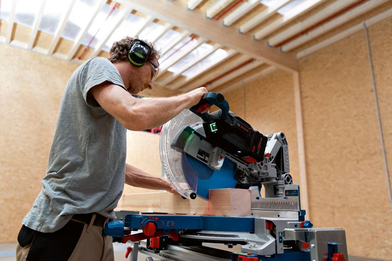 A person wearing safety equipment uses a cordless mitre saw to cut wood indoors.