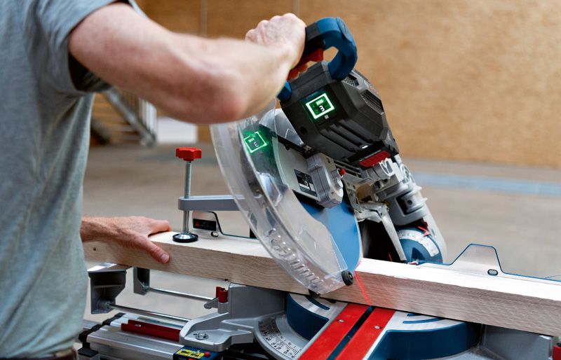 GCM 18V-305 GDC Person operates a cordless mitre saw to cut a wooden plank on a workshop table.