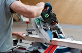 A person operates a cordless mitre saw to cut a wooden board indoors.