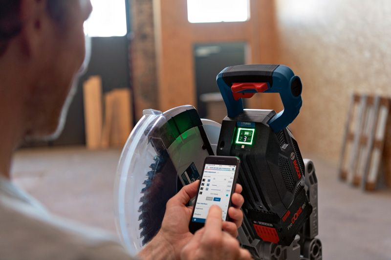 GCM 18V-305 GDC A person adjusts a cordless mitre saw with a smartphone in an indoor workshop.