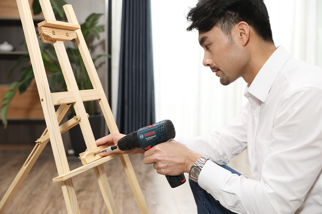 Person uses a power drill to assemble a wooden easel indoors.