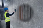 A person wearing safety equipment uses a detector on a concrete wall to identify hidden pipes.