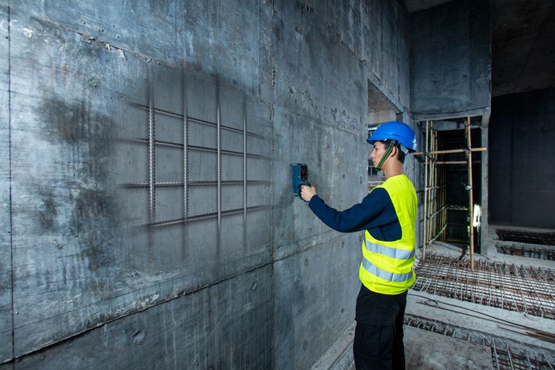 A person wearing safety equipment scans a concrete wall with a detector to locate rebar.