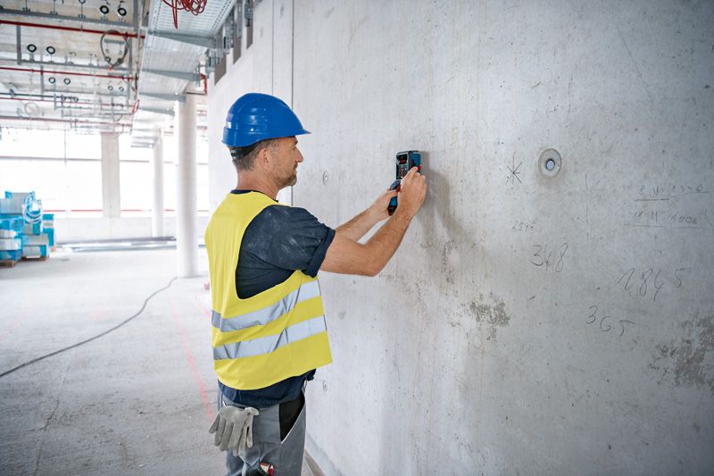 A person wearing safety equipment scans a concrete wall with a detector device.