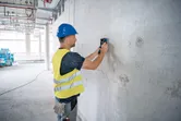 A person wearing safety equipment scans a concrete wall with a detector device.