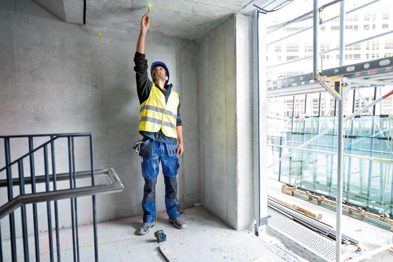 A person wearing safety equipment uses a point laser to mark the ceiling in a concrete room.