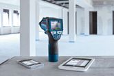 Thermo detector with digital display stands on a table next to two tablets in an unfinished room.