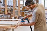 A person wearing safety equipment sands a wooden frame with a cordless random orbit sander.