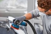 A person wearing safety equipment sands a car part with a cordless random orbit sander.