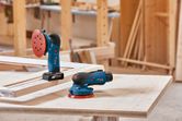 Cordless random orbit sanders placed on a wooden workbench in a carpentry workshop.