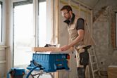 A person wearing safety equipment cuts wood with a cordless table saw in a renovation setting.