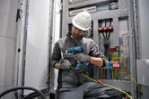 A person wearing safety equipment uses a cordless heat gun on electrical wiring.
