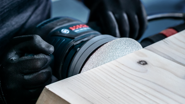 EXPERT M480 Sanding Net for Random Orbital Sanders 125 mm, G 80 5-pc Worker wearing safety equipment sanding a wooden plank with a power sander.