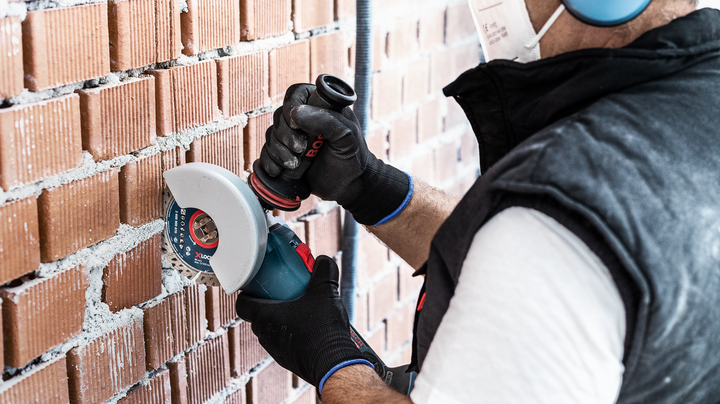 Person wearing safety equipment cuts into a brick wall with a power tool.