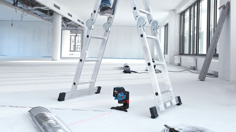 Laser leveling tool projecting lines on a floor beneath a person standing on a ladder.