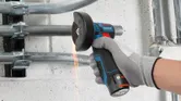 A person wearing safety equipment uses a cordless angle grinder to cut metal pipes.